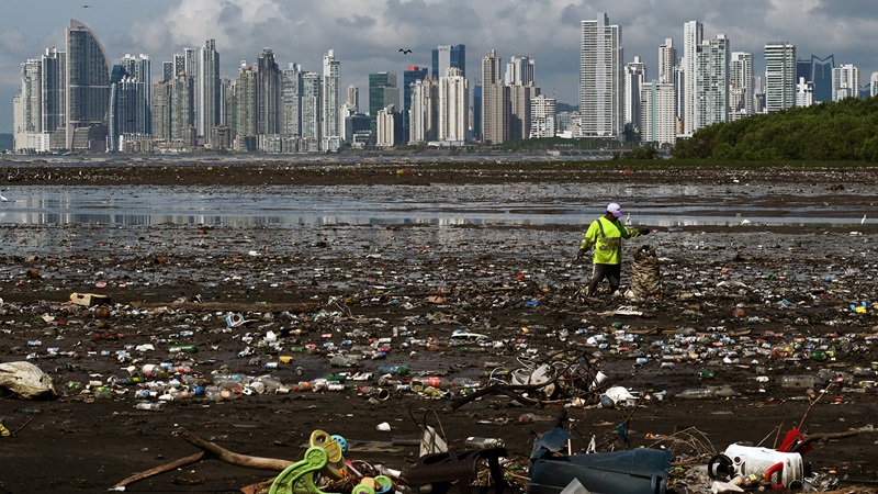 A person wearing a yellow jacket and white hard hat stands on a rubbish-strewn beach with a city skyline in the distance. 