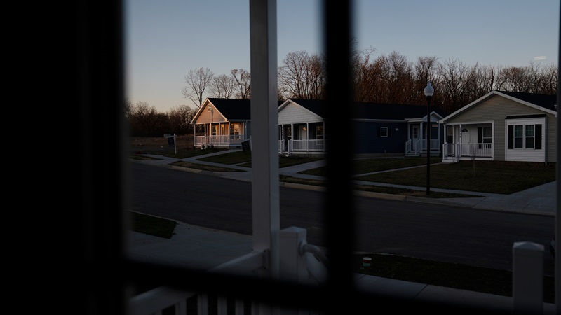 Three houses with pitched roofs, front porches, and front yards sit next to each other. They are viewed from the porch of a house across the street. 