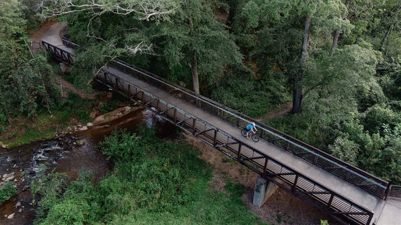 An overhead view shows a cyclist riding across a bridge built over a flowing river. The bridge and trees are surrounded by mature green trees and bushes. 